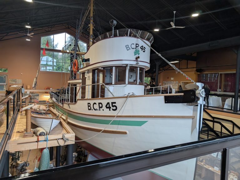 A large white boat labeled "B.C.P. 45" is displayed indoors on supports, with a small rowboat alongside it and various nautical items nearby. The space has high ceilings and windows in the background.
