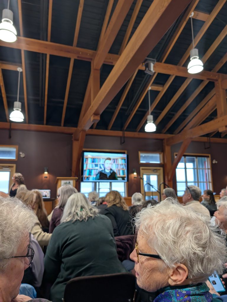 People seated in a room with wooden beams, watching a large screen showing a person speaking in front of bookshelves. Natural light comes through the windows.