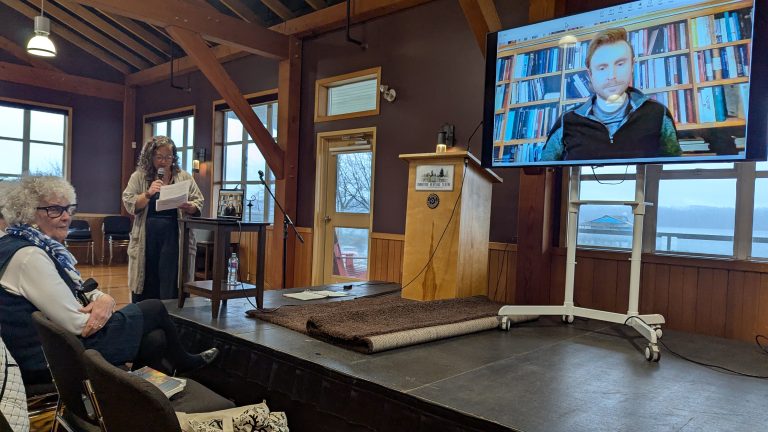 A woman speaks at a podium in a wooden room while an audience listens; a man appears on a large screen via video call, sitting in front of bookshelves.