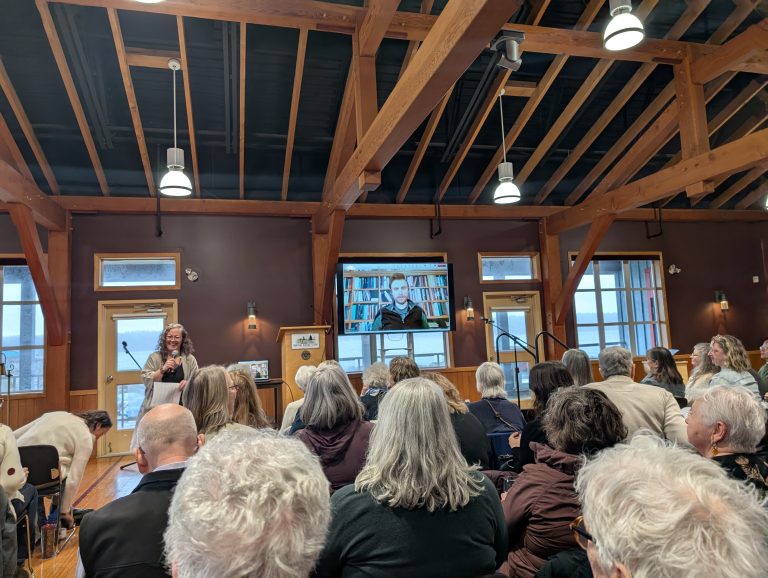 Audience seated in a wooden-beamed hall, listening to a woman speaking at the front; a large screen shows a person speaking via video call.