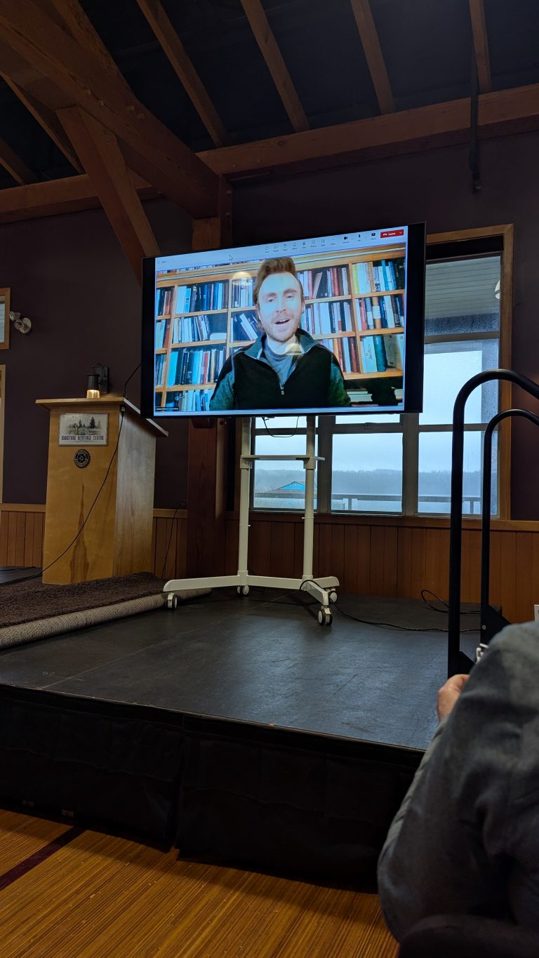 A large screen on a stage shows a man speaking on a video call, with bookshelves in the background. There is a podium and window nearby, and part of an audience member’s arm is visible in the foreground.
