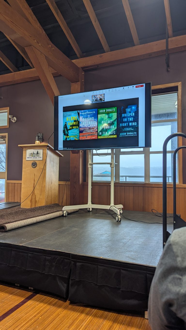 A TV screen displays book covers during a presentation on a stage in a wooden room with large windows. Nearby is a podium and a rolled-up carpet, with lake and tree views outside.