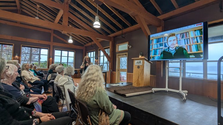 A group of people in a wood-beamed room watches a woman speak at a podium while a man appears on a large video screen beside her. Windows show trees and water outside.