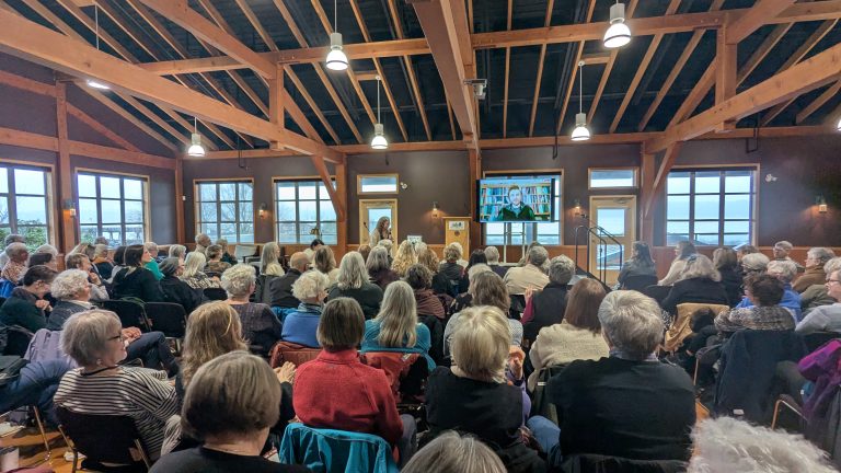 A large group of people sit in a spacious wooden hall, attentively watching a speaker at a podium and a screen displaying another person speaking via video call.