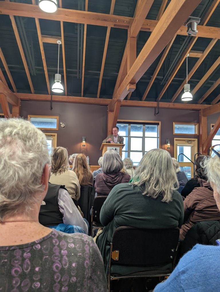 A person speaks at a podium to an audience seated in a room with wooden beams, large windows, and natural light.