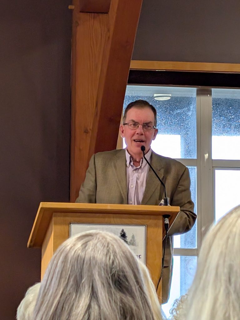 A man wearing glasses and a brown blazer speaks at a podium with a microphone. There are people with gray hair in the audience and a window behind him.