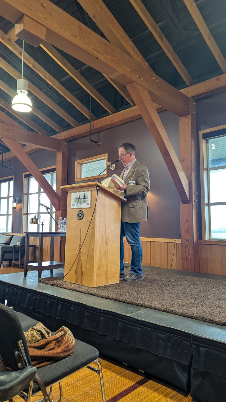 A person stands at a wooden podium, reading from a book in a room with wooden beams, windows, and a small audience.