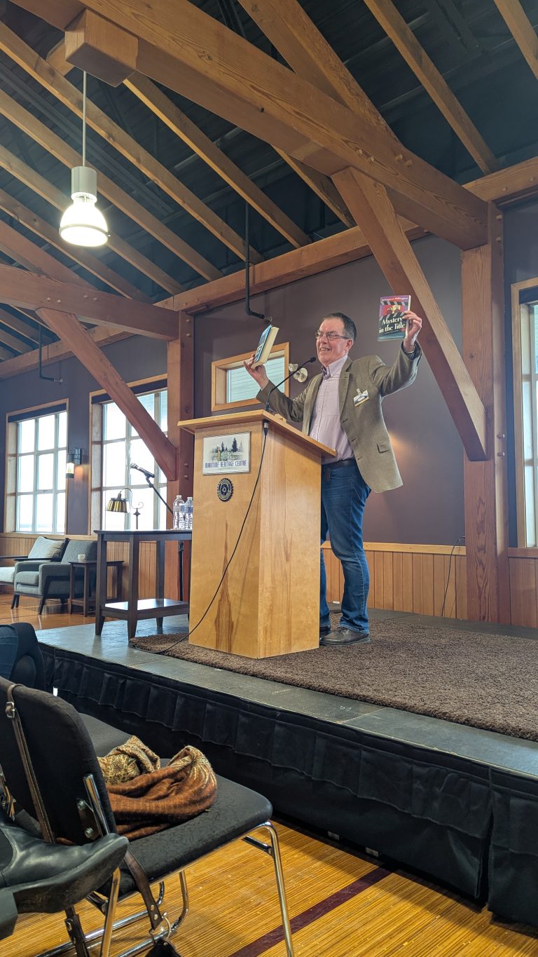A man stands on a stage behind a wooden podium, smiling and holding up two books, one in each hand, in a sunlit room with exposed beams and wooden walls.