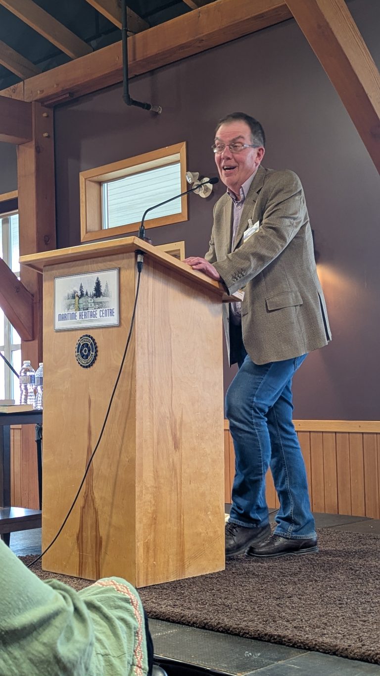 A man in a brown blazer and jeans stands at a wooden podium, smiling and speaking in a rustic room with exposed beams.