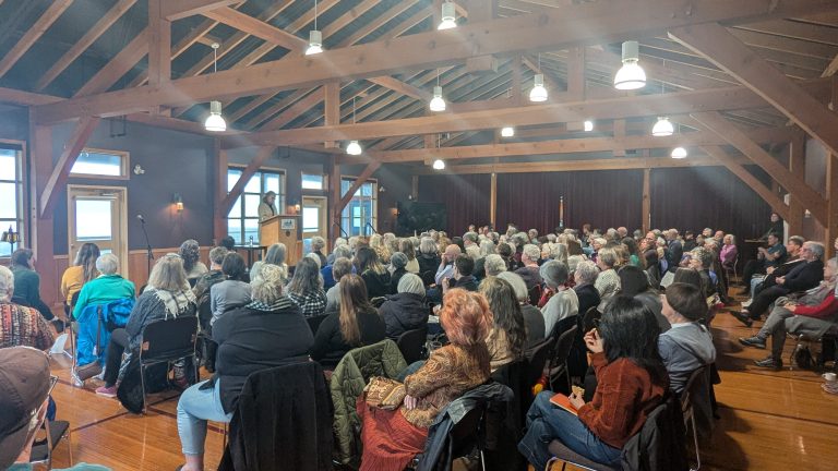 A large group of people sit in rows of chairs facing a speaker at a podium in a wooden, high-ceilinged room with exposed beams and large windows.
