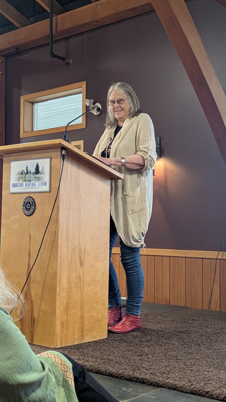 An older woman stands at a podium labeled “Maritime Heritage Centre,” smiling as she speaks to an audience in a wood-paneled room.