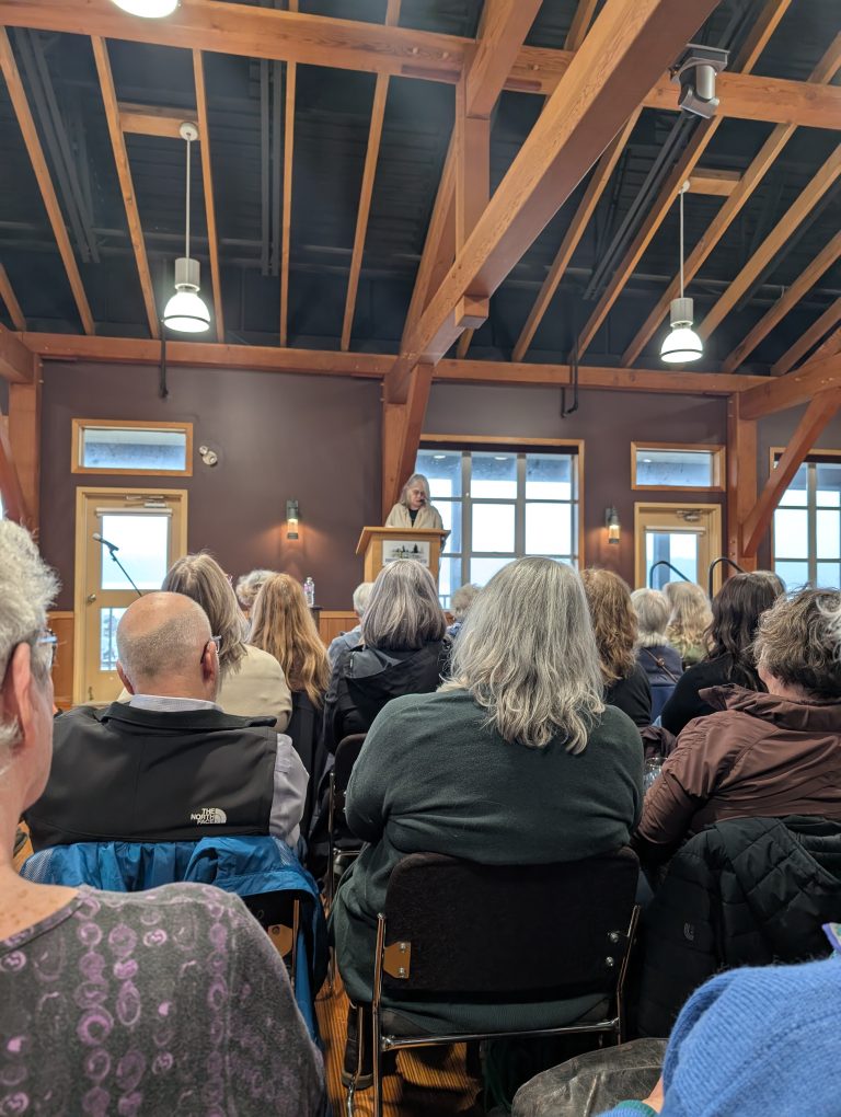 A speaker stands at a podium addressing an audience seated in a room with wooden beams, large windows, and hanging lights. The crowd listens attentively.