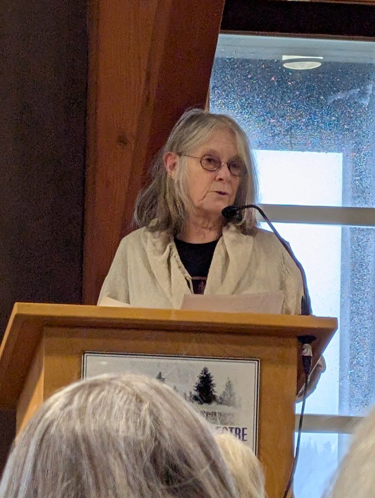 An older woman with gray hair and glasses is speaking at a podium indoors, holding papers. She stands before a microphone, with a window and audience visible in the background.
