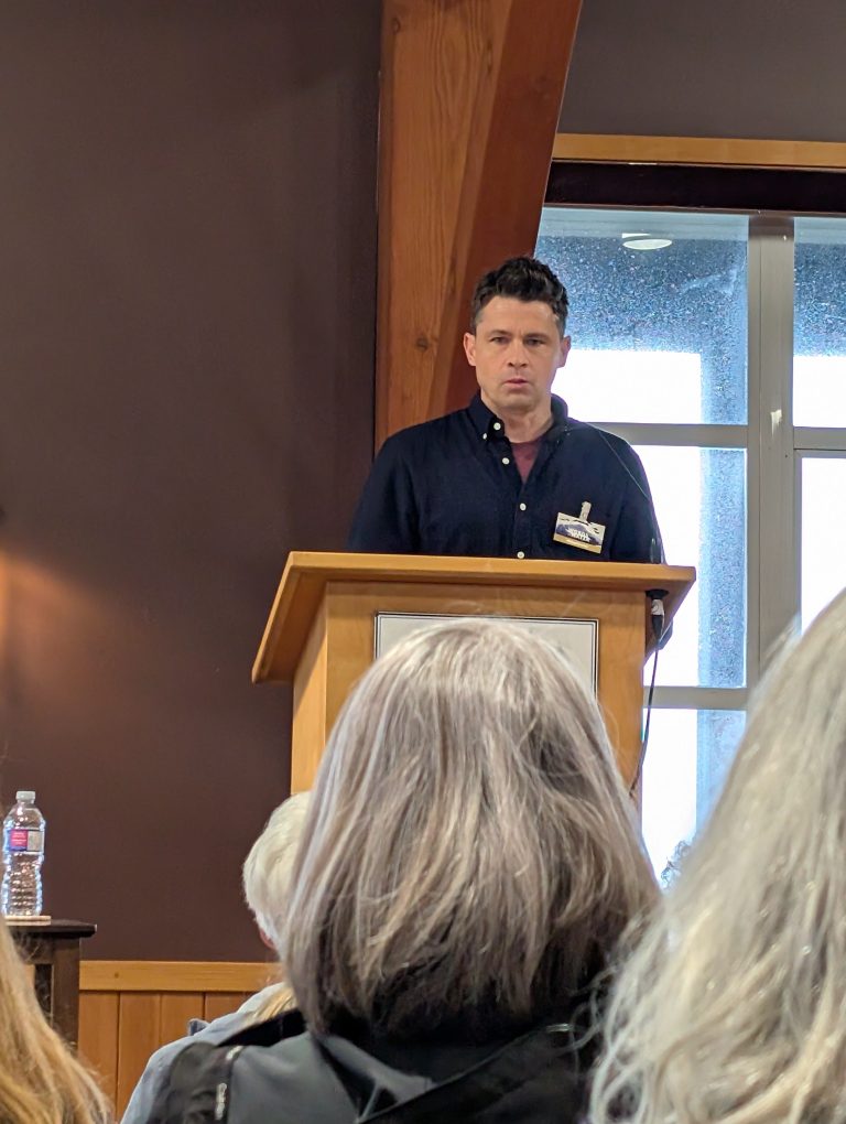 A man stands at a wooden podium speaking to an audience in a room with dark walls and windows behind him. People with gray hair are seated and listening.