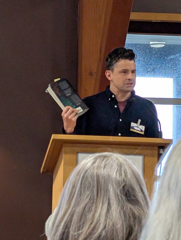 A man stands at a wooden podium, holding up two books while speaking to an audience. He wears a dark shirt and name tag. The image is taken from behind some seated people.