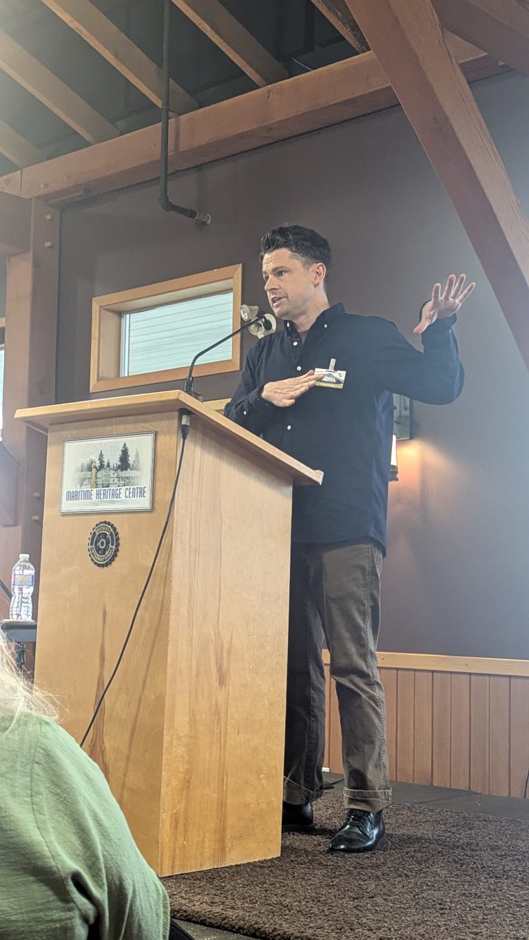 A man speaks at a wooden podium labeled "Maritime Heritage Centre," gesturing with one hand raised and the other on his chest. He stands indoors with exposed beams and a window in the background.