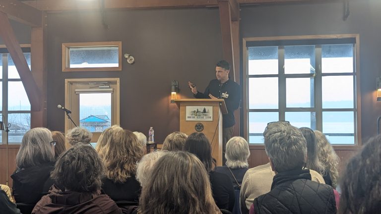 A man speaks at a podium to a seated audience in a room with large windows and wooden beams. A microphone and water bottle sit nearby. The scene overlooks water outside.
