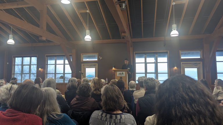 A speaker addresses an audience seated in a wooden hall with large windows and exposed beams. The room is filled with natural light and attendees are focused on the podium.