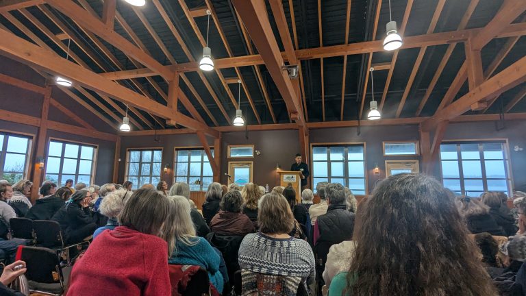 A large group of people sits facing a speaker at a podium in a wooden-beamed hall with large windows; the audience listens attentively.