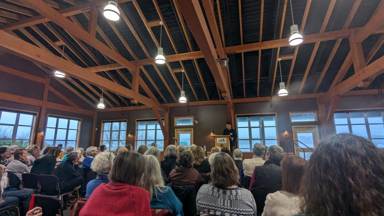A large audience sits in a wooden-beamed hall, attentively listening to a speaker at a podium in front of large windows. Bright ceiling lights illuminate the room.