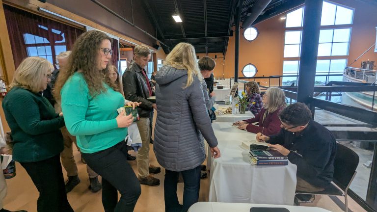 People wait in line at a book signing event. Two authors sit at a table, signing books, while attendees stand and hold books, waiting for their turn. The setting is indoors with large windows in the background.