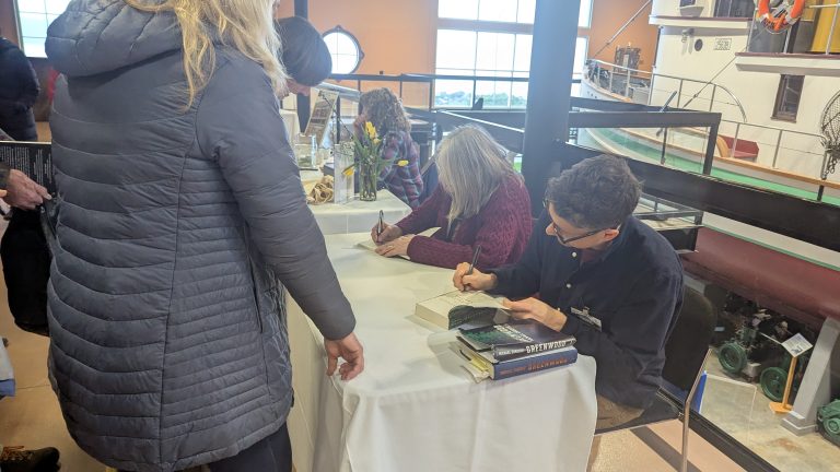 Two authors sit at a table signing books for attendees at an indoor event. Stacks of books and flowers are on the table, and a large boat is visible in the background. People stand in line to meet the authors.