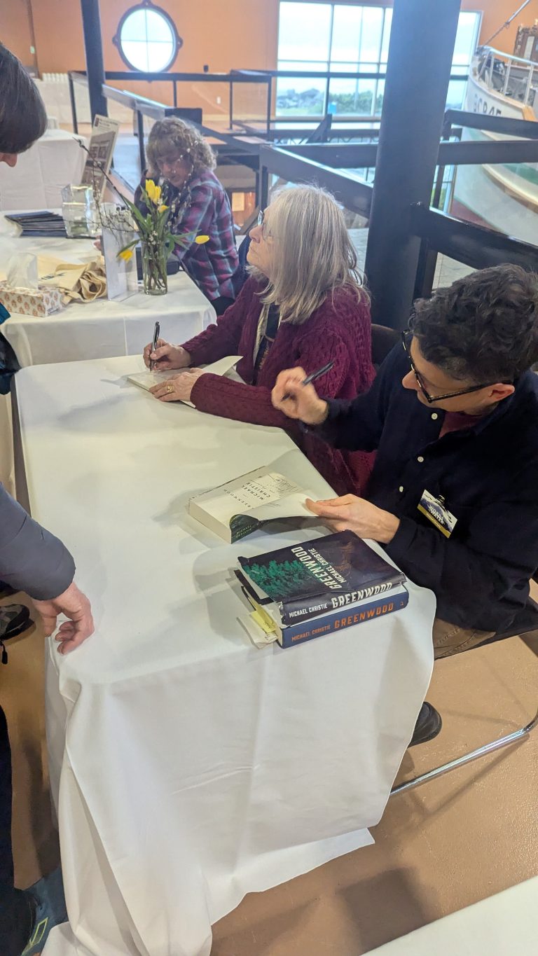 Two people sit at a table signing books at an indoor event. One person writes in a book while the other prepares books for signing. A vase of yellow flowers and more books are on the table.