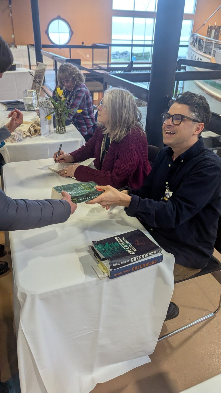 Two authors sit at a table signing books for attendees at an indoor event. One author is handing a signed book to a person, while the other is signing a book. Stacks of books and a vase of flowers are on the table.
