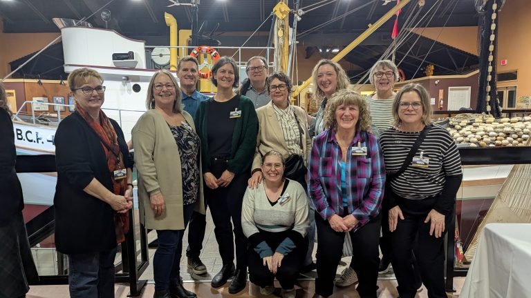 A group of twelve adults smiles for a photo inside a maritime-themed museum, standing and kneeling in front of a boat display and nautical decor.