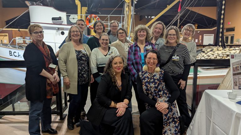 A group of people smiling and posing together indoors at a maritime-themed museum, with a boat and display cases in the background.