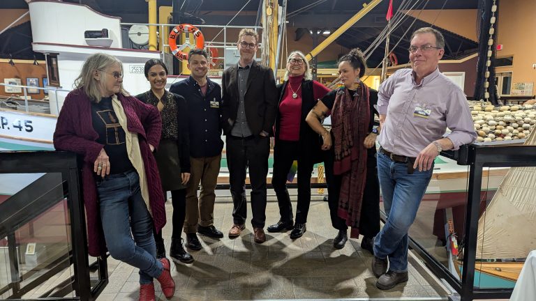 Seven people pose and smile together indoors near a boat display, with a net of buoys and maritime artifacts in the background. Most are casually dressed, standing side by side.
