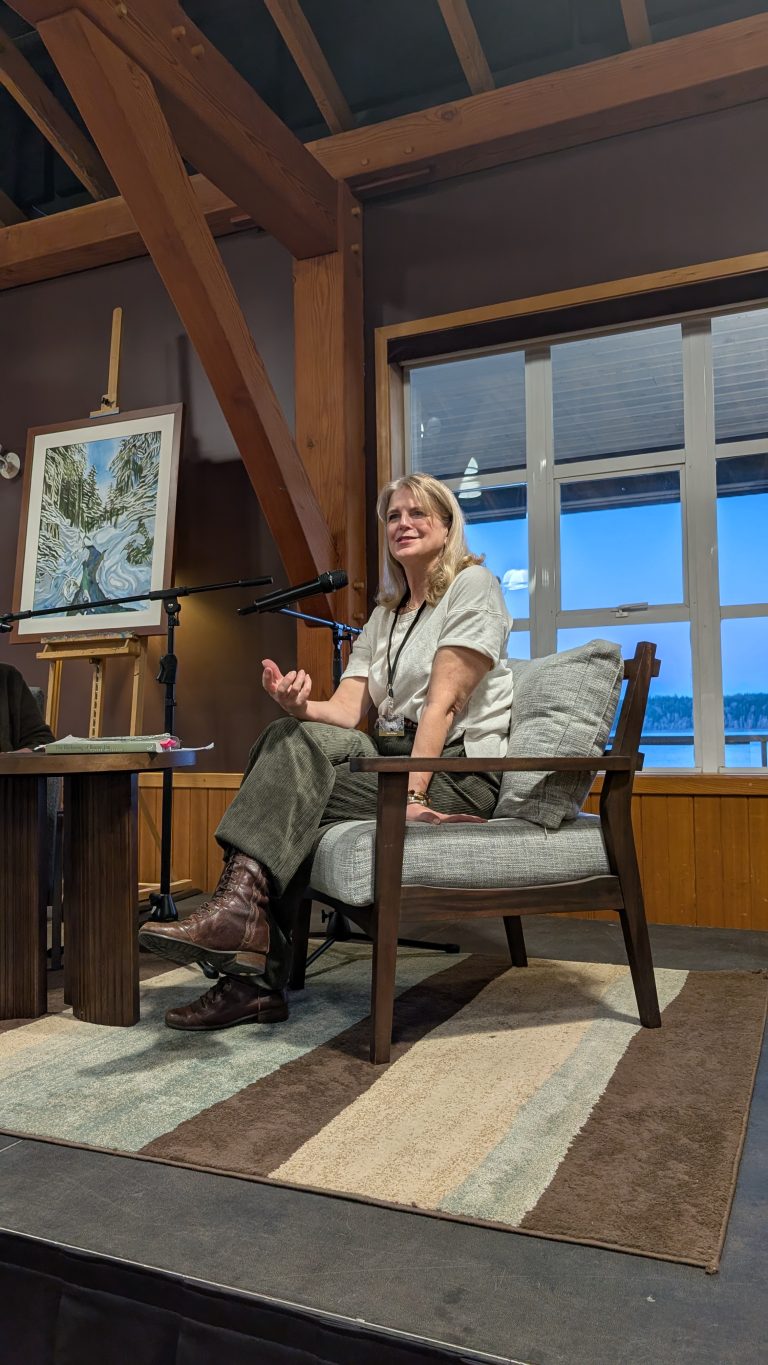 A woman sits in a chair on a stage, speaking, with a large window and a painting on an easel behind her. She gestures with one hand, and the setting appears to be a cozy indoor event space.