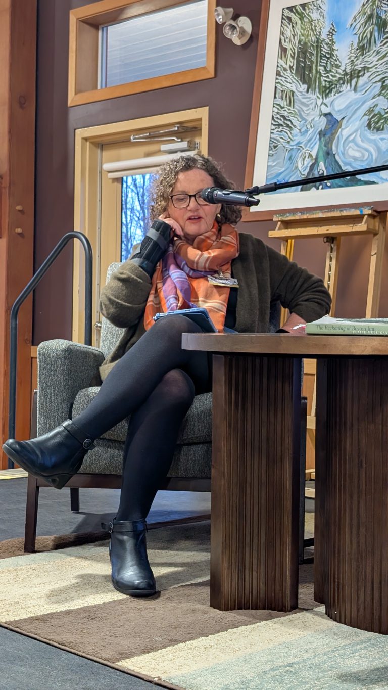 A woman with curly hair, glasses, and an orange scarf sits in an armchair, speaking into a microphone. She is holding a pen and paper, with books on the table beside her. A snowy landscape painting is in the background.