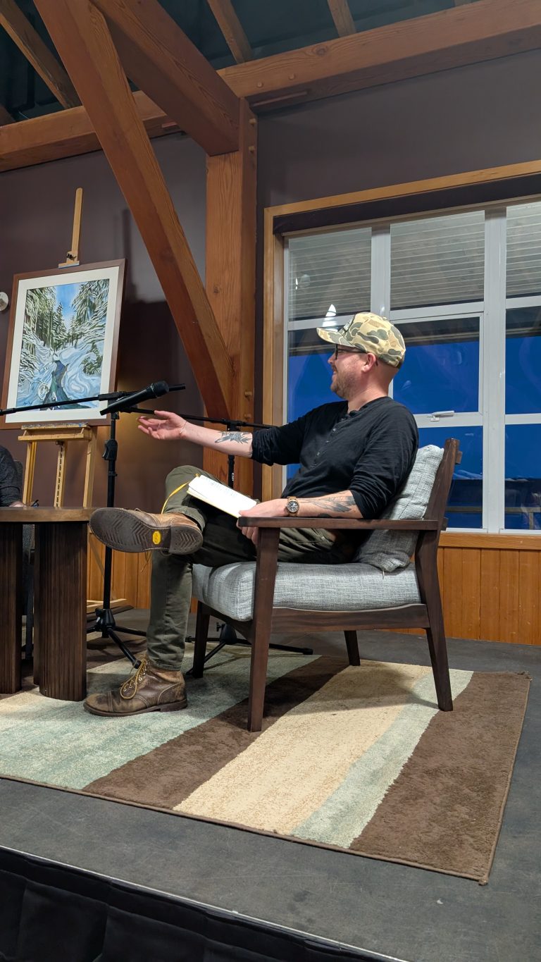 A man in a cap and glasses sits on a chair, holding a notebook and gesturing while speaking into a microphone. A painting on an easel is next to him, with wooden beams and large windows in the background.