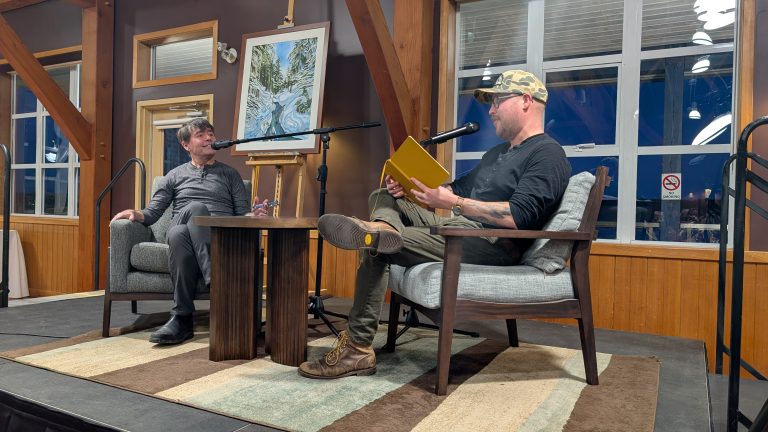 Two men sit facing each other on stage in armchairs, one holding a yellow book and speaking into a microphone. A painting on an easel sits between them. Wooden beams, windows, and a rug are visible in the room.