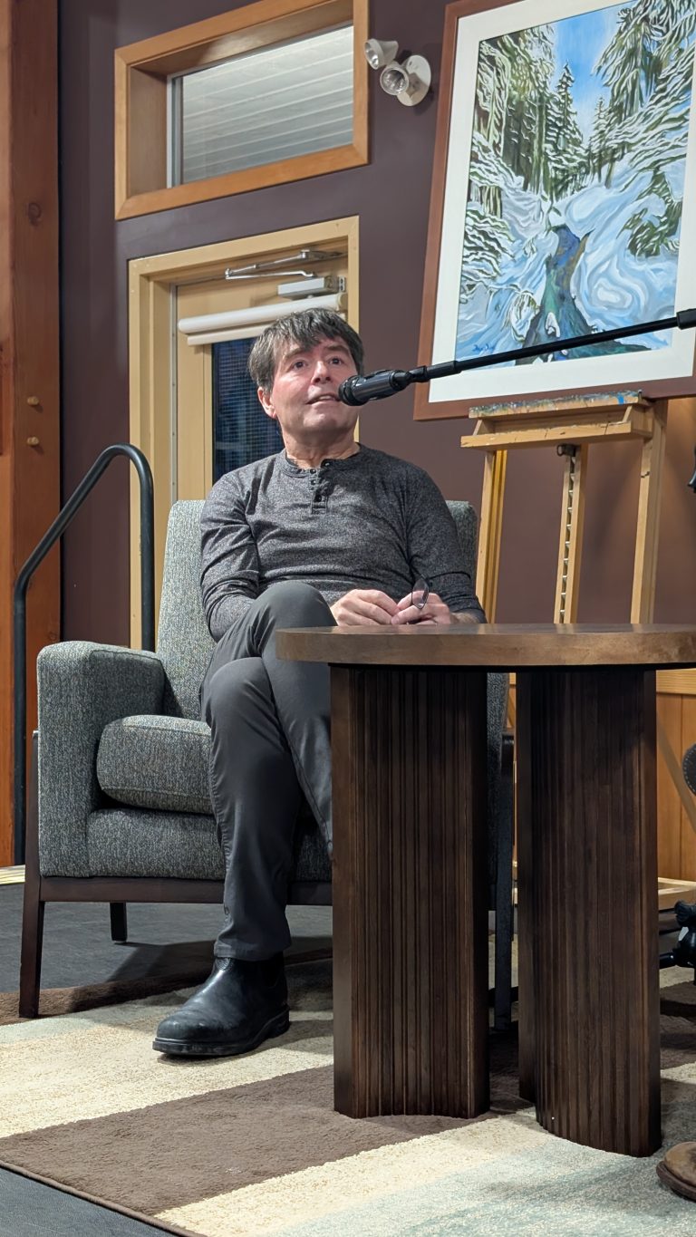 A person sits on a gray armchair, speaking into a microphone. They are on a stage with a painting of a snowy forest scene on an easel behind them. A wooden table is in the foreground.