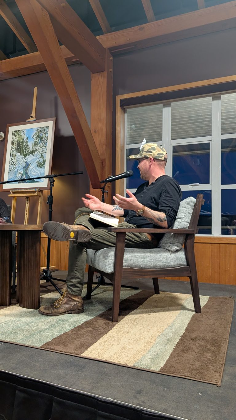 A man in a camouflage cap sits on a chair with a book, gesturing while speaking into a microphone. A painting on an easel and another microphone are beside him. The room has wooden beams and a striped rug.
