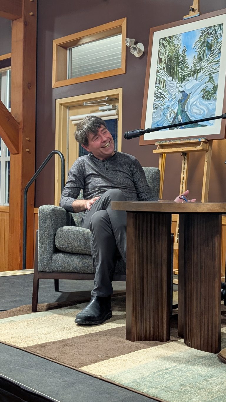 A person sits smiling in a gray chair on stage next to a wooden table, with a microphone and a winter landscape painting on an easel nearby. The background features wooden beams and windows.