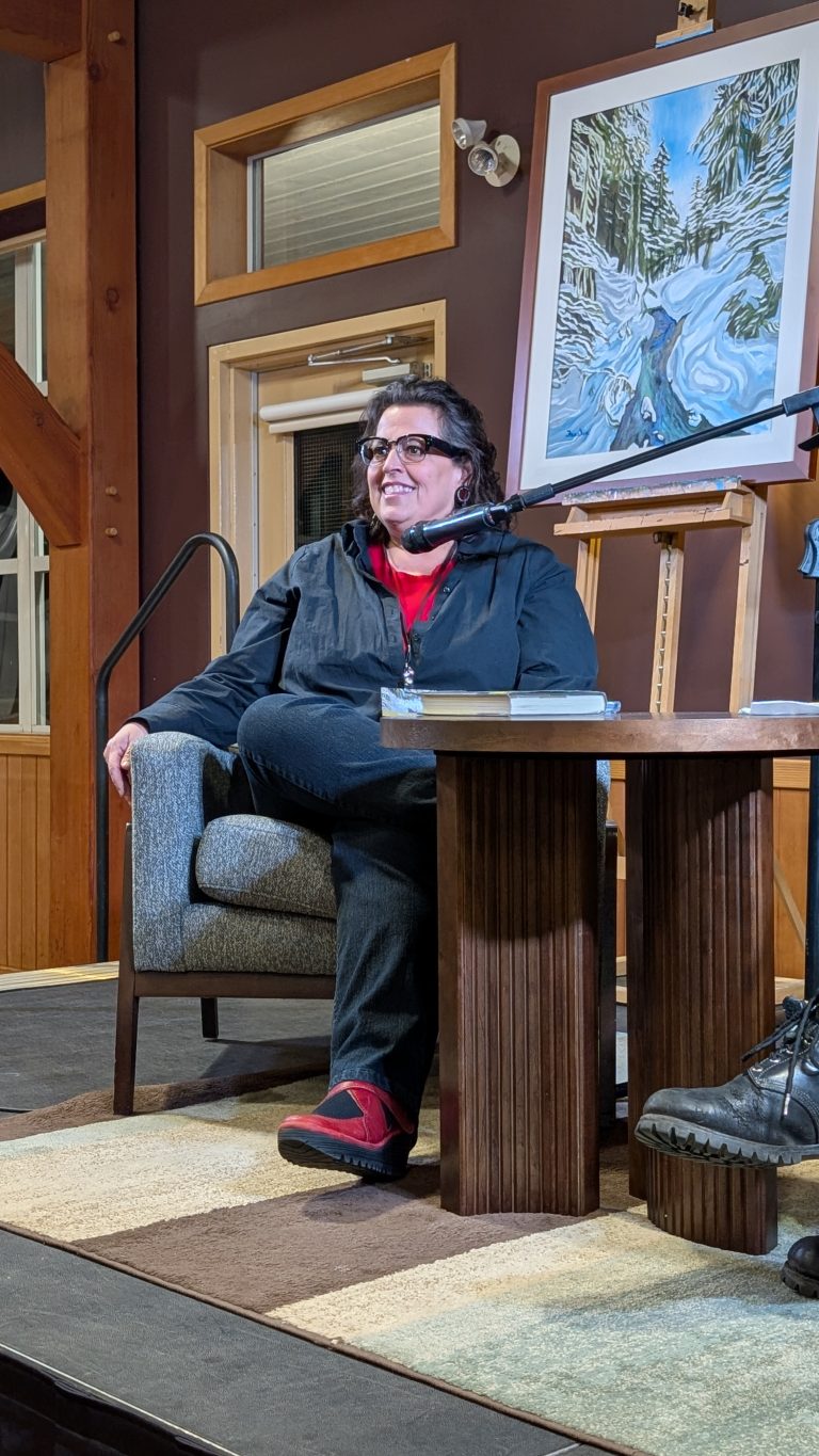 A person with glasses sits on a gray chair, smiling, next to a table with books. A painting of a snowy landscape is displayed on an easel behind them. The setting appears to be an indoor event or talk.