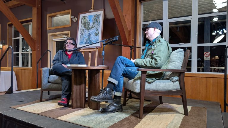 Two people sit in armchairs on a small stage, facing each other with microphones. A painting on an easel stands behind them. Large windows and wooden beams are visible in the background.