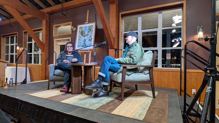 Two people sit on a stage having a conversation. They are seated in armchairs with a small table between them. A painting on an easel is behind them, and the room has large windows and wooden beams.