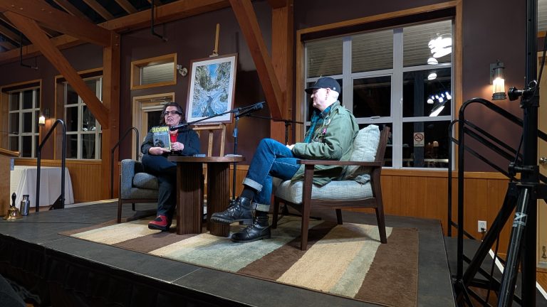 Two people sit on a stage having a conversation, with a painting on an easel behind them. Wooden beams, windows, and lights fill the cozy room. Both are seated in armchairs on a small rug.
