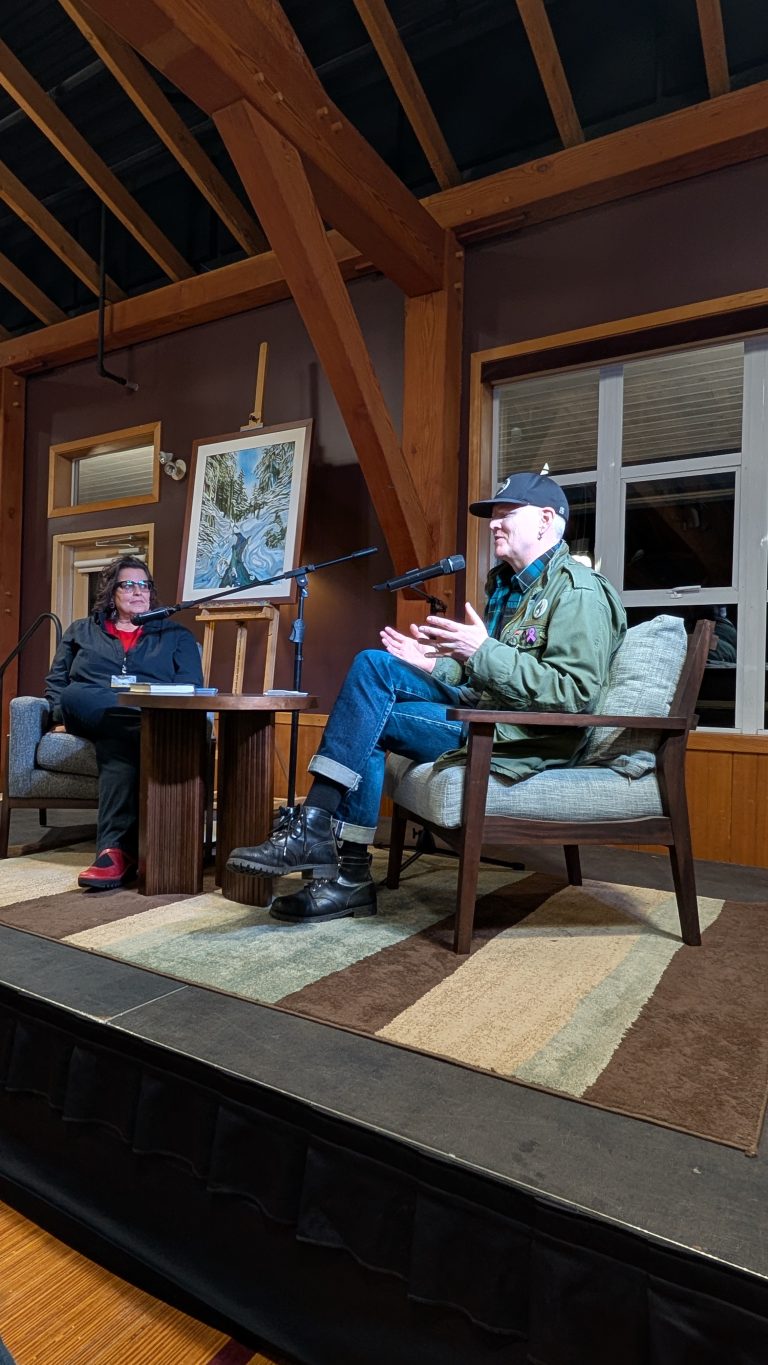 Two people sit on stage having a conversation. One person speaks into a microphone, gesturing with their hands. The room has wooden beams, a large window, and art on the wall.