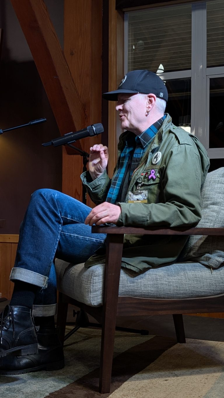 A person wearing a cap, green jacket, jeans, and boots sits on a chair, speaking into a microphone. The setting appears to be indoors with wooden beams and a window in the background.