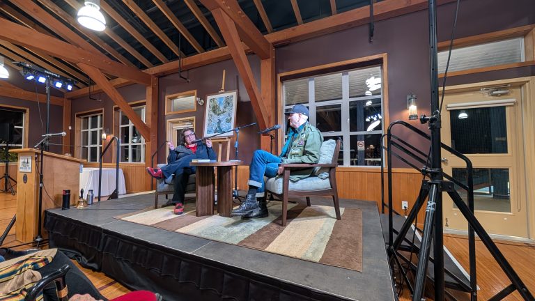 Two people sit and talk on a stage in a rustic room with wooden beams, large windows, and indoor lighting. A table with drinks is between them, and audio equipment is visible.