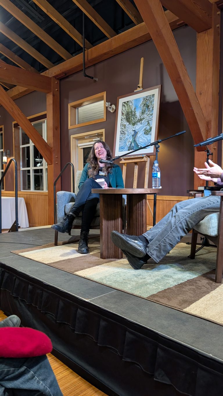 A woman sits on stage, smiling, next to a table with a water bottle. Another person's legs are visible. Wooden beams and a painting of a river and trees are in the background.