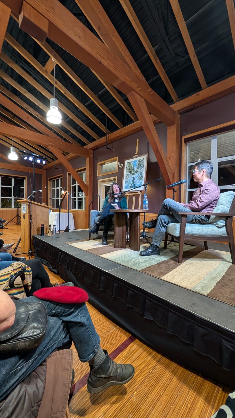 Two people sit on stage having a conversation in a wooden-beamed room. Audience members watch from the foreground. A large painting rests on an easel between the speakers.