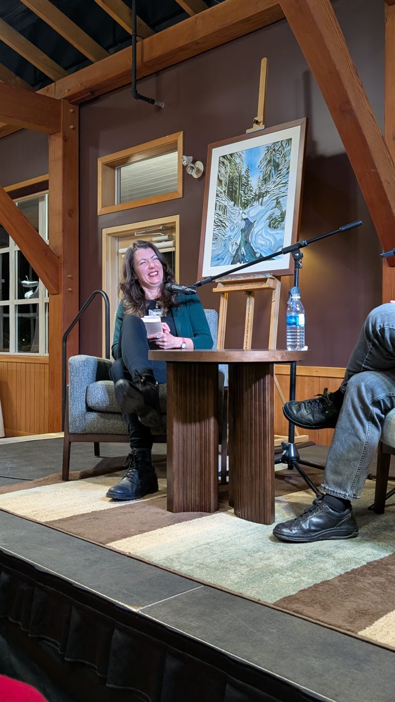 A woman sits on stage smiling, holding a microphone, next to a man. There's a painting, water bottle, and two tables between them. The setting is a cozy indoor space with wooden beams.