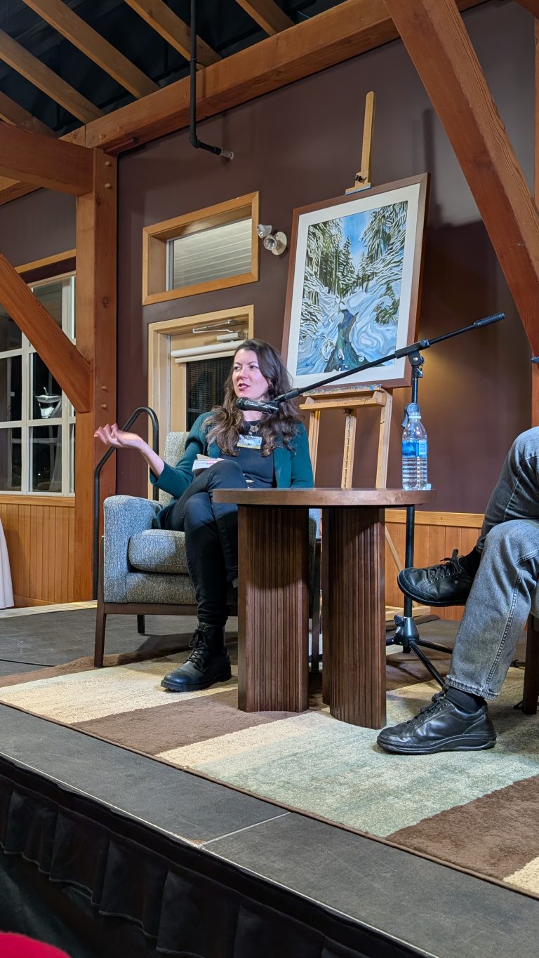 A woman sits onstage in an armchair, gesturing as she speaks into a microphone. A table, water bottle, and partial view of another person are visible. Behind her is a painting of a snowy landscape on an easel.