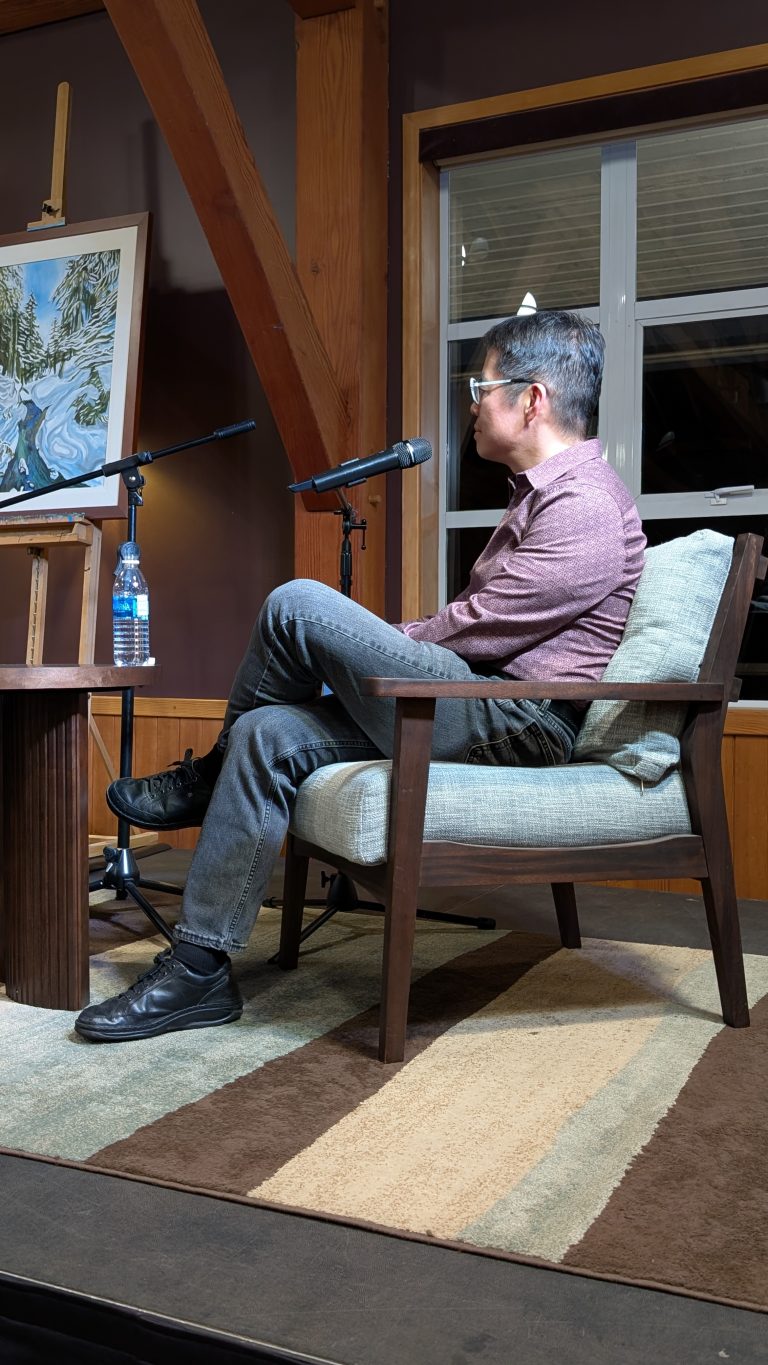 A man sits on a chair with legs crossed, facing two microphones. A water bottle and a painting are on a small table near him. He is indoors, next to a window and wooden beams.